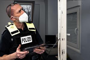 Das Bild zeigt einen Polizisten mit einem Tablet, der neben einem Fensterrahmen mit offensichtlichen Einbruchsspuren steht. Der Polizist inspiziert den Fensterrahmen, um die Einbruchsspuren anschließend mit Hilfe des Tablets zu dokumentieren.