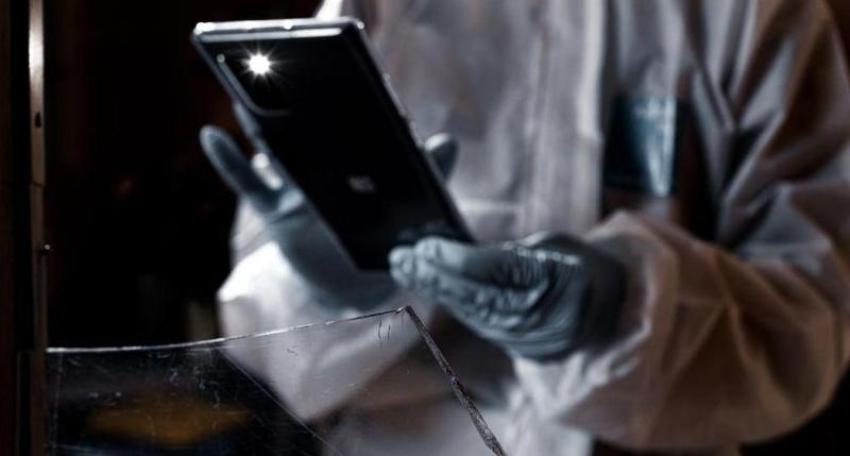 Das Bild zeigt eine eingeschlagene Fensterscheibe. Im Hintergrund ist eine spurensichernde Person zu sehen, die mit dem Smartphone ein Foto der Fensterscheibe aufnimmt.